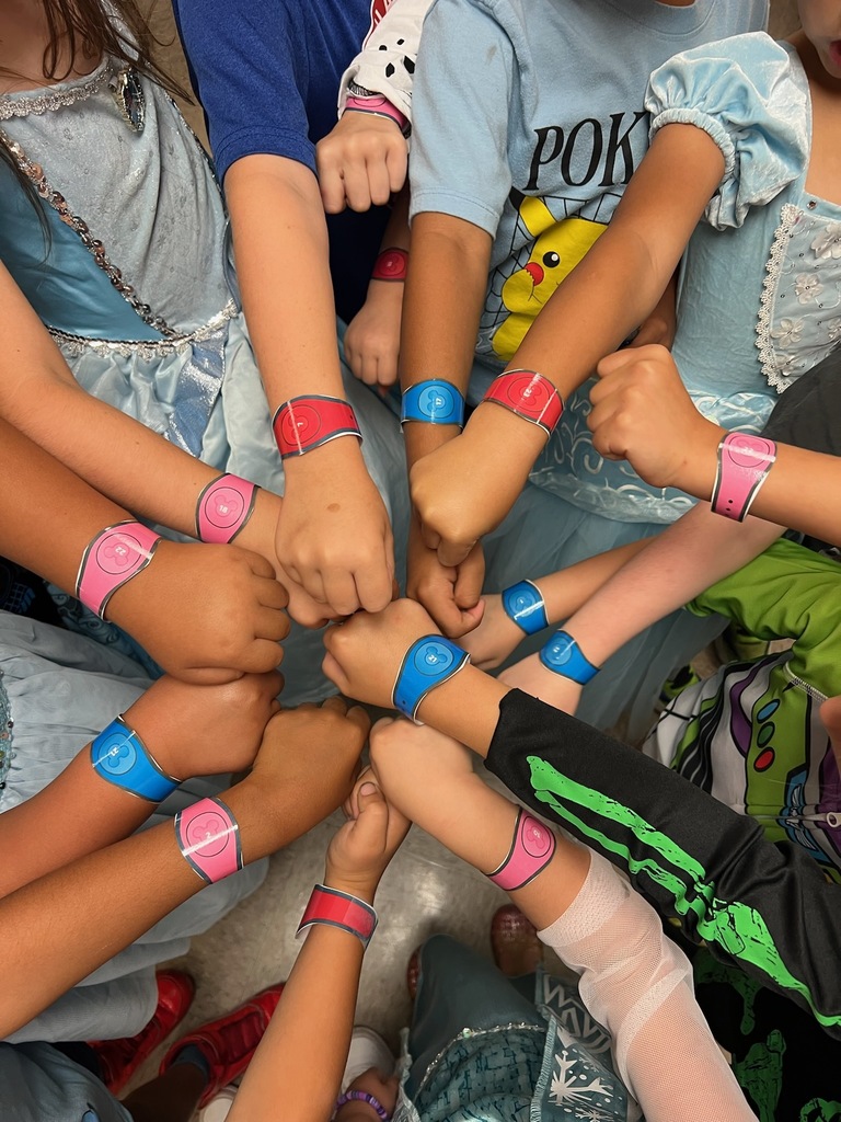 Children gather, their arms forming a circle. Some wear colorful bracelets. They are seated on the floor.