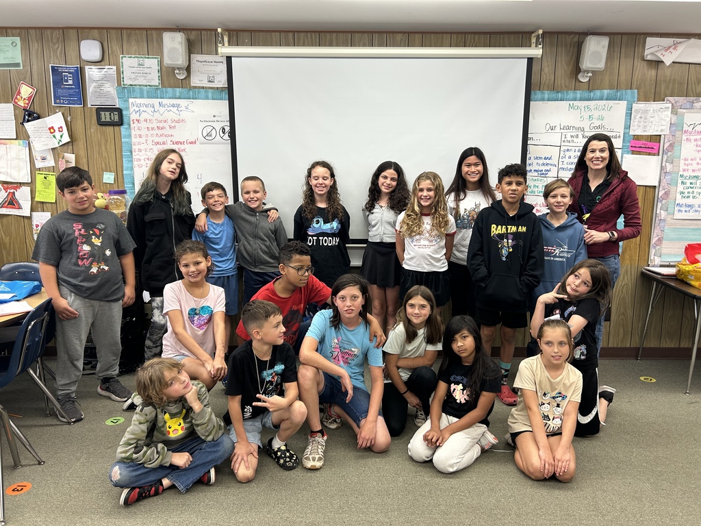 Students of varying ages sit and stand in a classroom. Some sit on the floor and others on chairs. A teacher stands among them.