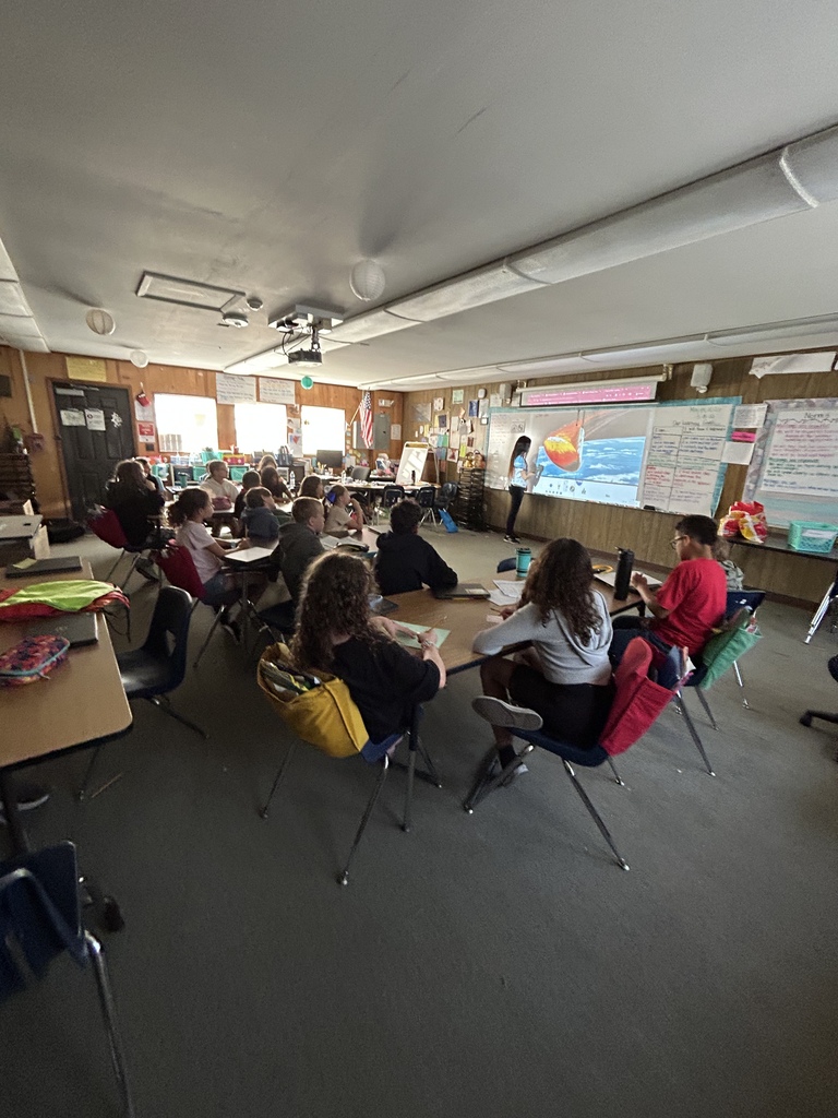 A classroom with students seated at tables. Whiteboards and a projector screen are on the wall.