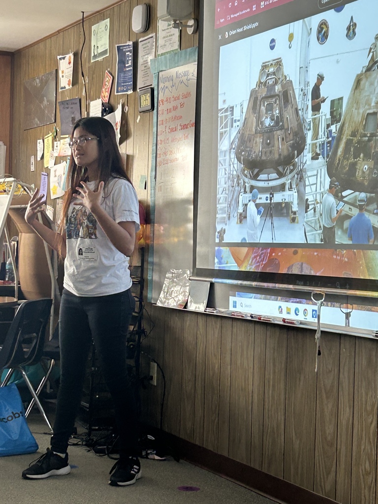 A person stands in front of a large screen with a spacecraft image, wearing glasses and sneakers.