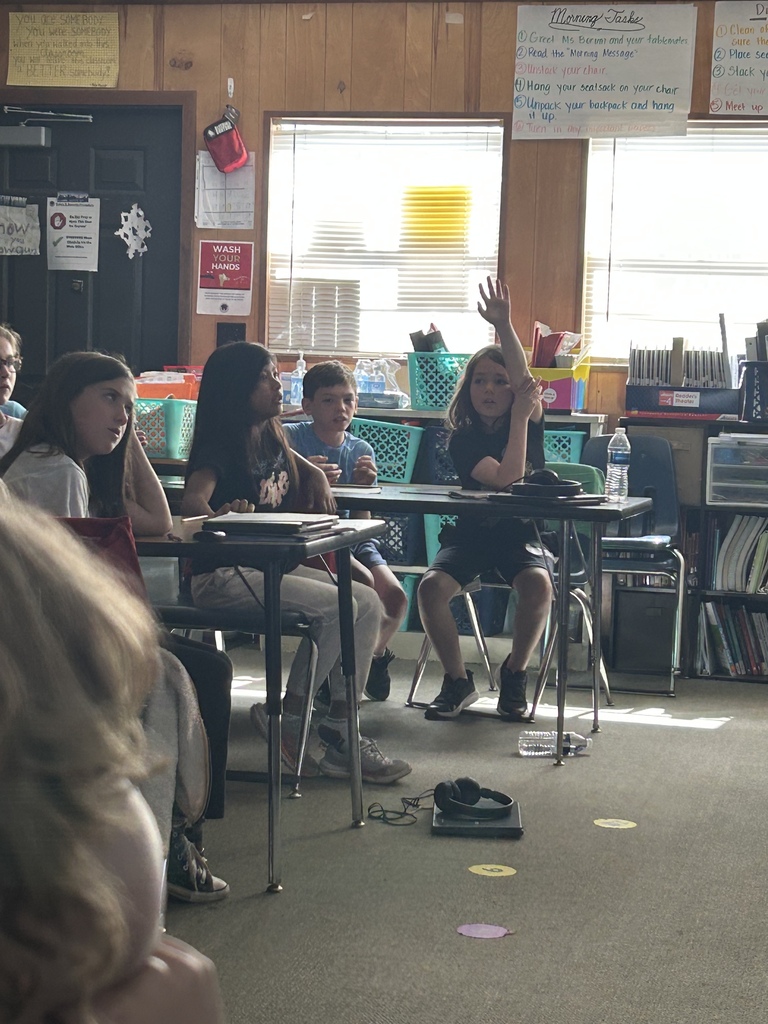 A classroom with several students seated around desks. One student raises their hand. Shelves with books and storage bins are in the background.