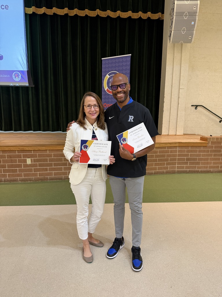Two people stand in front of a stage holding certificates. A woman wears a white suit and glasses, a man wears a black shirt and sneakers.