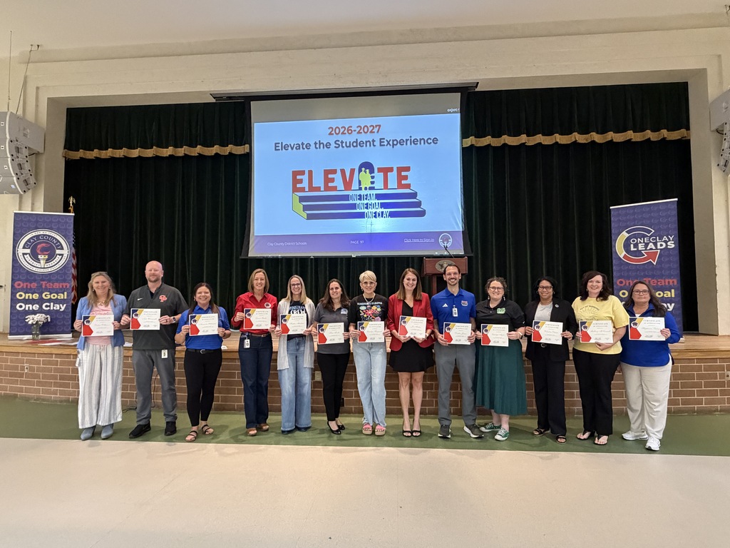 A group of people holds certificates in a room with banners and a projector screen displaying Elevate.