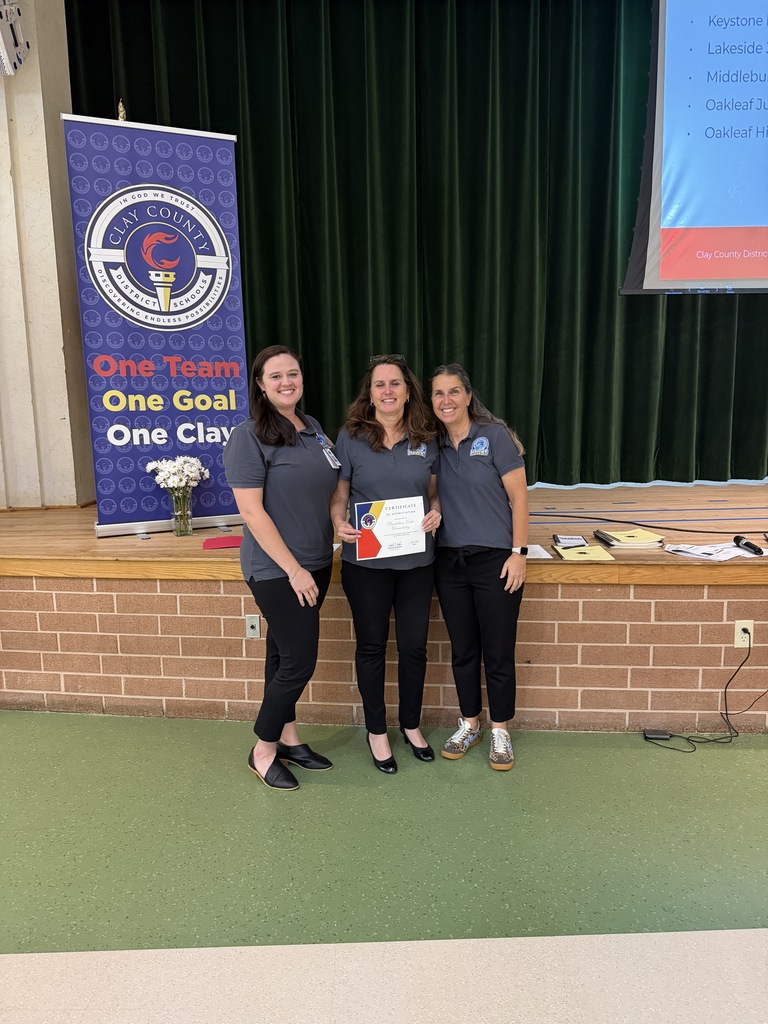 Three women stand before a brick wall, holding an award. They wear gray shirts and black pants. Behind them, a banner and a projector screen are visible.
