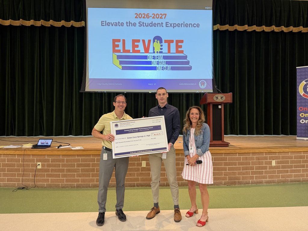 Three people stand in front of a stage holding a large check. Behind them is a screen showing "2026-2027 Elevate the Student Experience".
