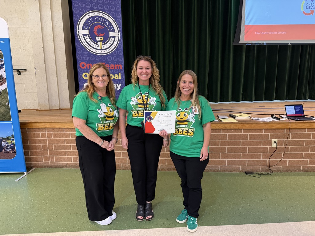 Three women in green T-shirts and black pants stand in a room with a brick wall, smiling and holding a certificate.