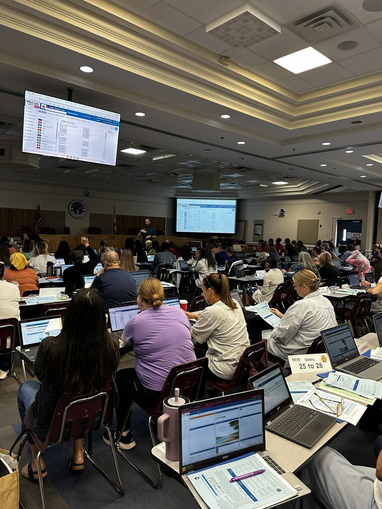 Large room where many people sit at tables with laptops. A screen is visible on the wall.