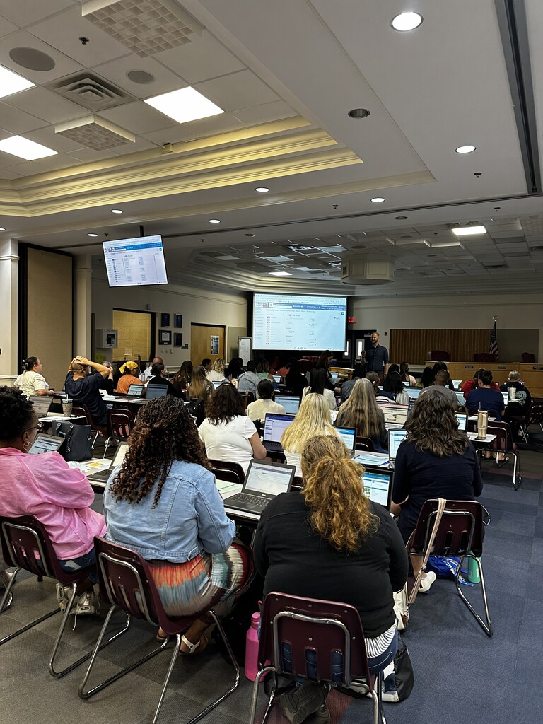 Large group in a room with chairs and laptops. A man stands with a screen behind him.