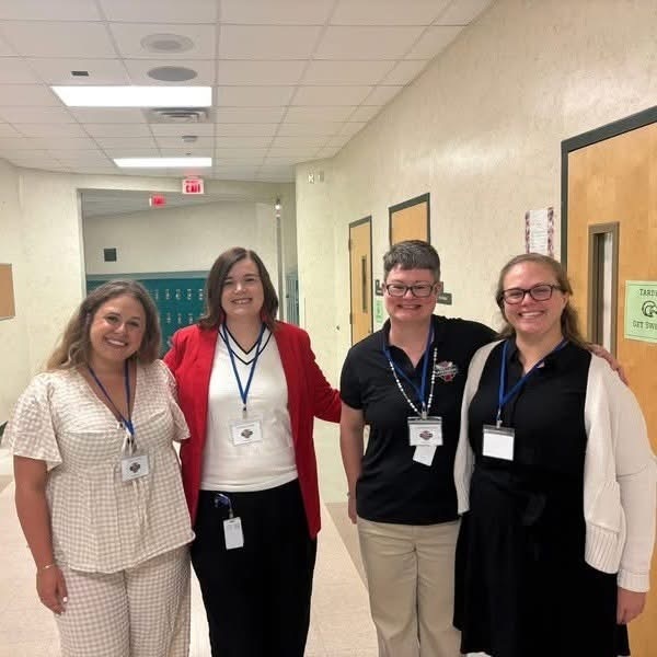 Four women stand in a hallway, wearing IDs. They have their arms around each other and smile.