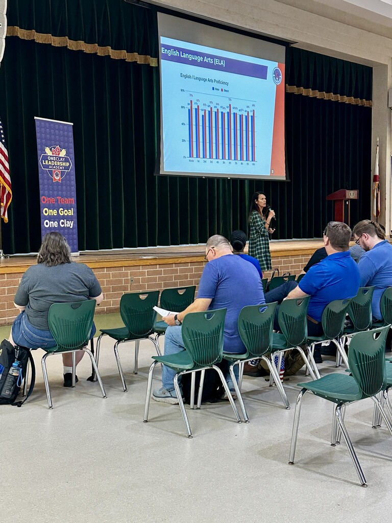 A woman at a podium gives a presentation with a screen behind her. Several people are seated in chairs, watching.