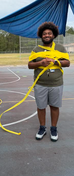 A person stands on a basketball court with yellow tape wrapped around their upper body, smiling and posing for a photo.