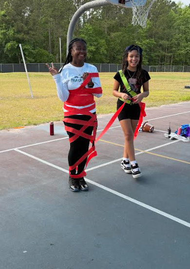 Two girls stand on a basketball court, one wearing a red and white shirt with red ribbons and the other a black shirt with a ribbon.