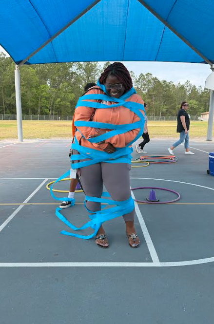Person stands on a basketball court with blue tape wrapped around them, surrounded by hula hoops. Trees in the background.
