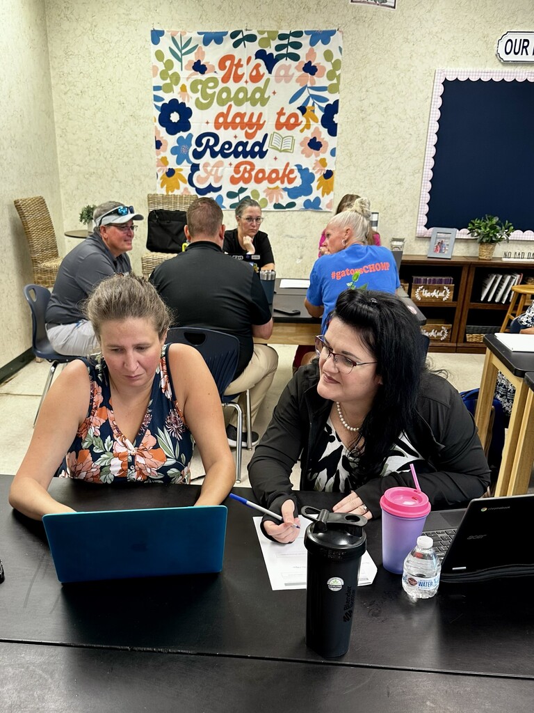Two women seated at a desk, each using a laptop, with one holding a pen and paper. A motivational sign hangs in the background.