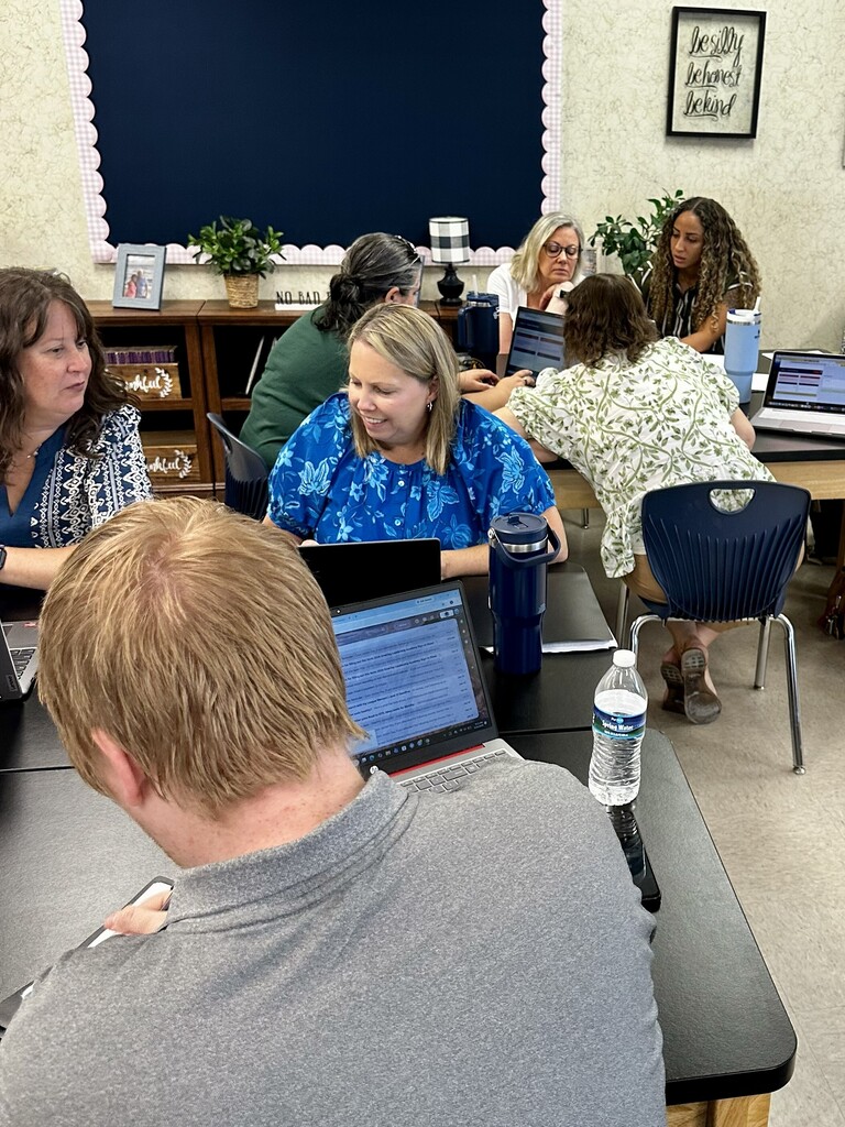 A group of people seated at desks, looking at laptops. A woman in a blue shirt is in front.