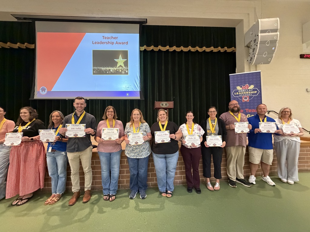 Nine people stand in front of a stage with certificates in their hands. A projector screen shows an award announcement.