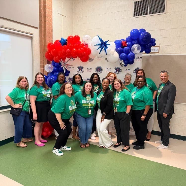 A group of people in green shirts stand close together in front of a red, white, and blue balloon arch.