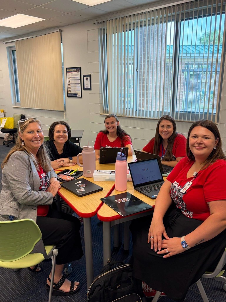 Five women sit at a table in a room with windows, laptops, and other items on the table.