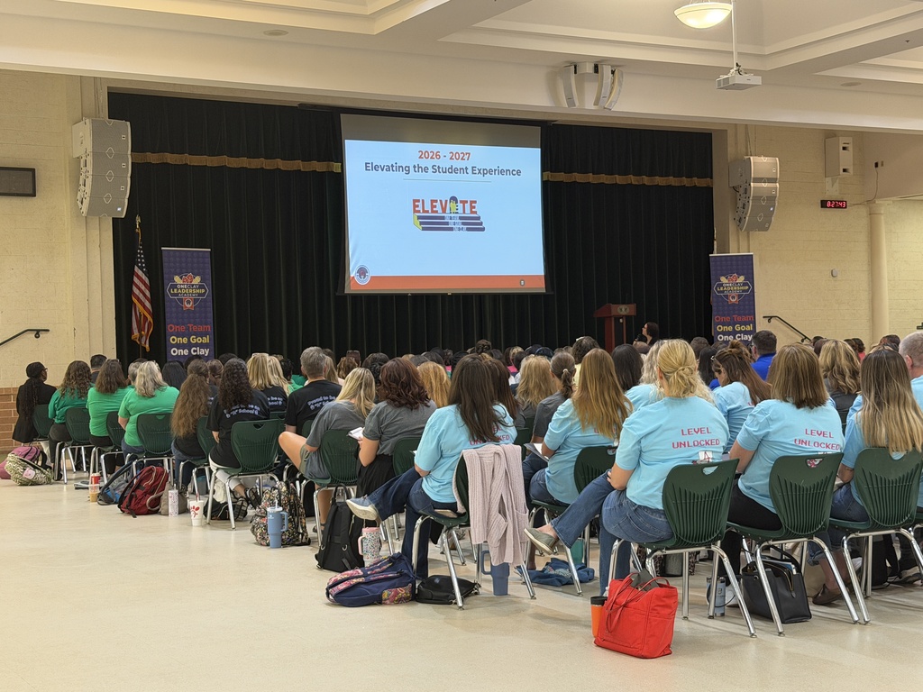 A room with a stage and screen shows a presentation to people seated in chairs, some wearing green shirts.
