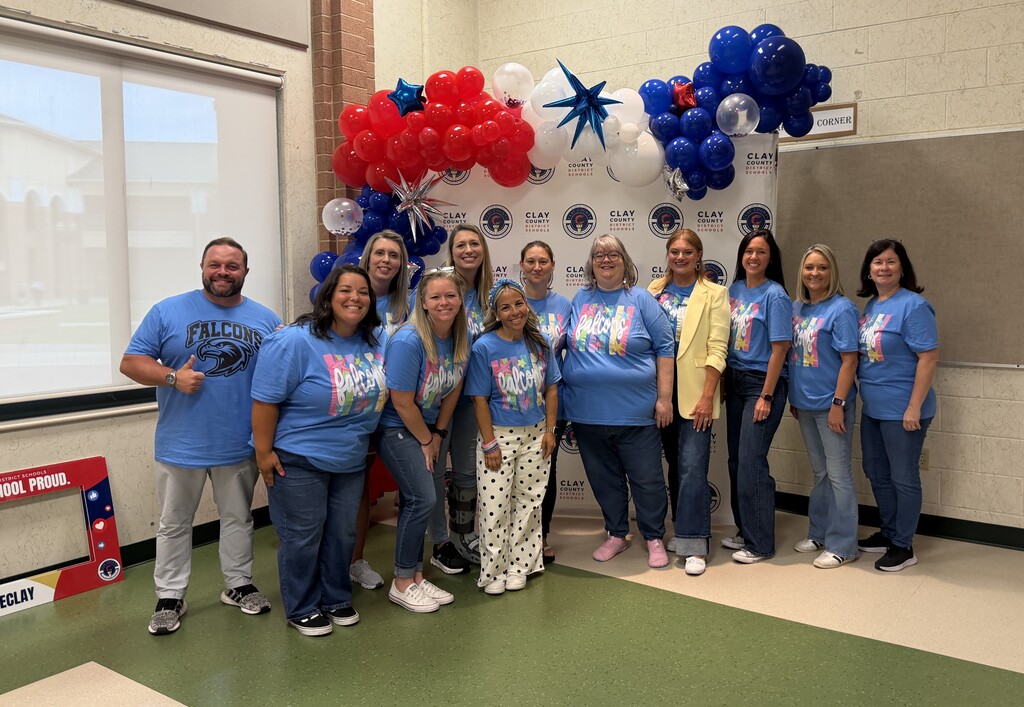 A group of adults in blue shirts stand in a room, with balloons and a banner in the background.