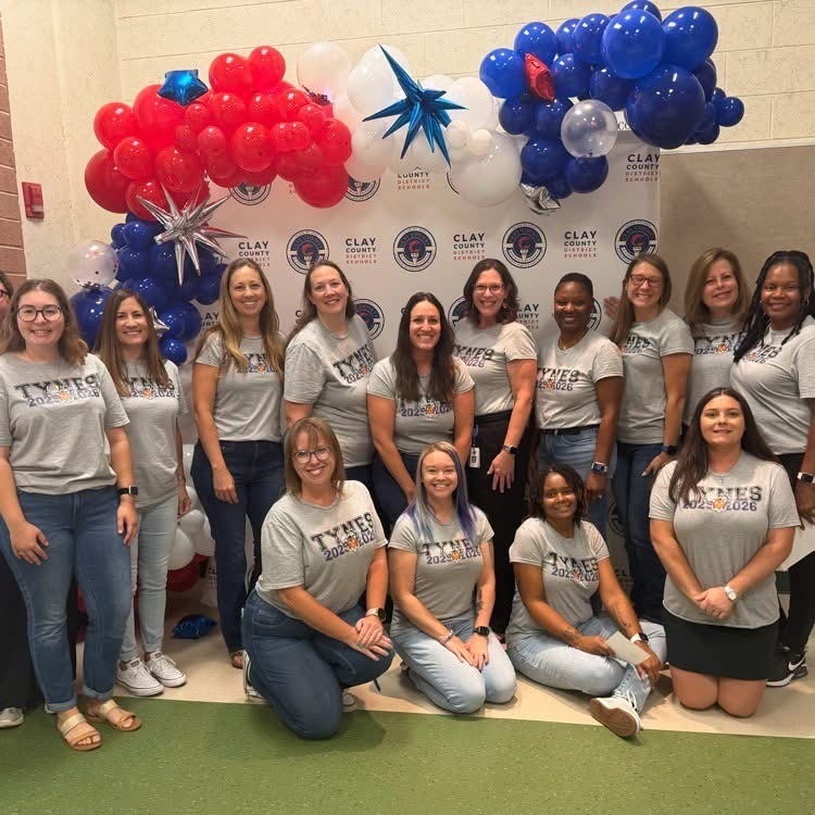 A group of women wearing gray t-shirts poses for a photo in front of a backdrop with balloons.