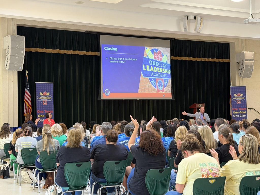 A room with a projector screen. A man speaks to an audience seated in chairs. Flags are visible.
