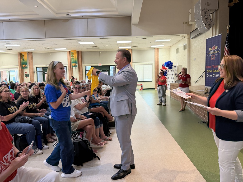 A man in a suit hands a ribbon to a woman in a blue shirt. They stand in a room with an audience seated on benches.