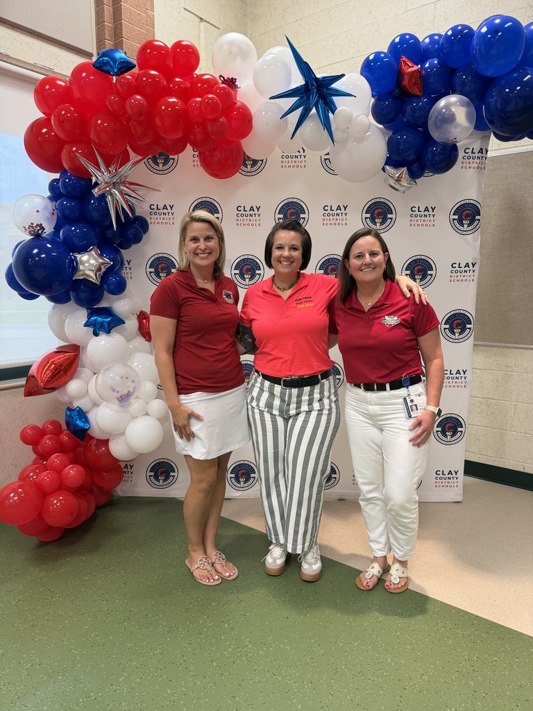 Three women in red shirts and white pants stand in front of a patriotic balloon arch.