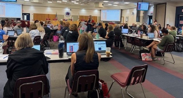 Large room with many people sitting at tables, each using laptops. Monitors on walls, ceiling lights, and carpeted floor.
