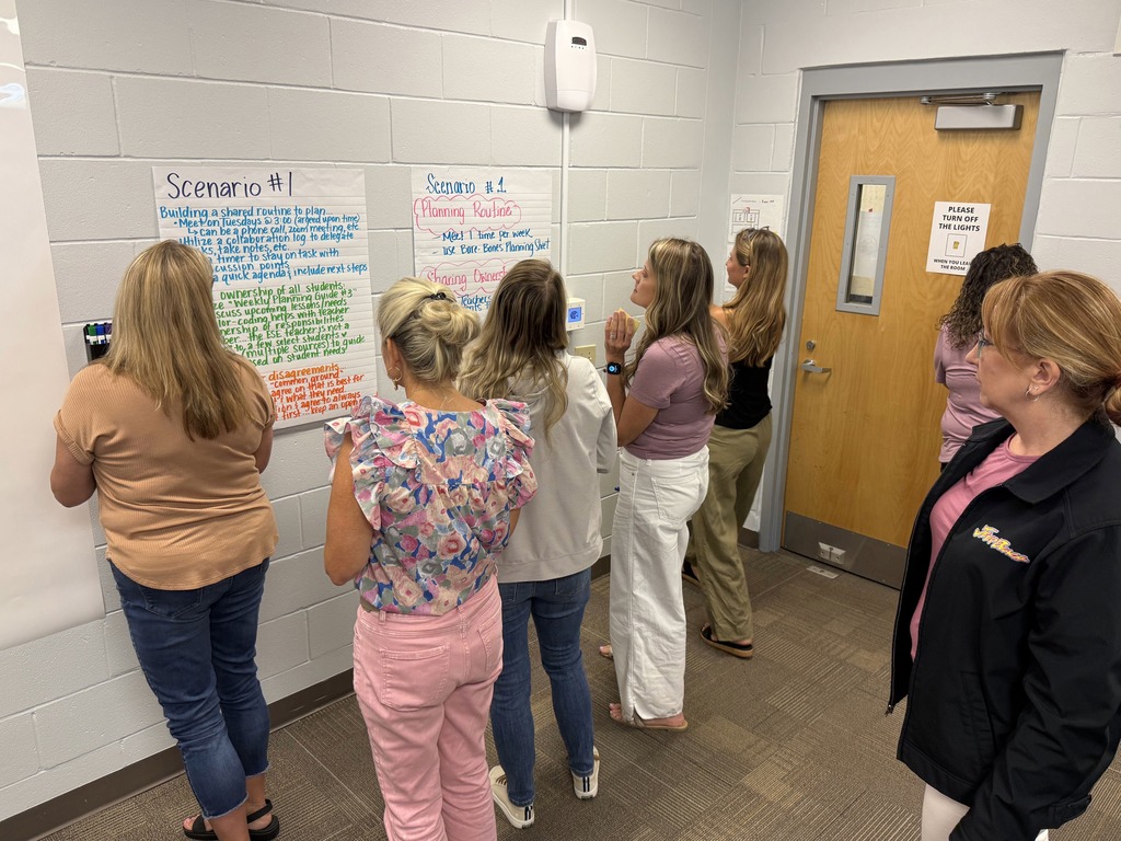 A group of women stands around a bulletin board, examining posted scenarios, in a room with a wooden door.
