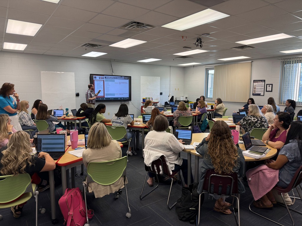A classroom with rows of people sitting at desks, engaged in an activity, with laptops and a projector screen at the front.