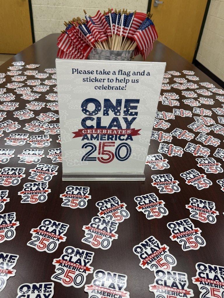 A table covered with stickers and flags. A sign says "One Clay Celebrates 250". There are red, white, and blue flags.