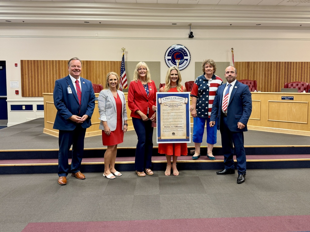 People stand in a council chamber. Two women hold a framed certificate. Two American flags are behind them.