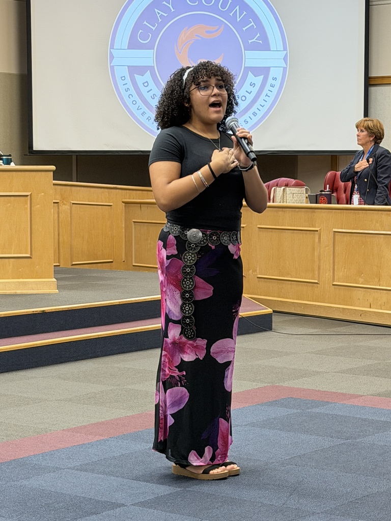 Woman in black top and floral skirt holds microphone in front of podium, with Clay County sign on screen behind.
