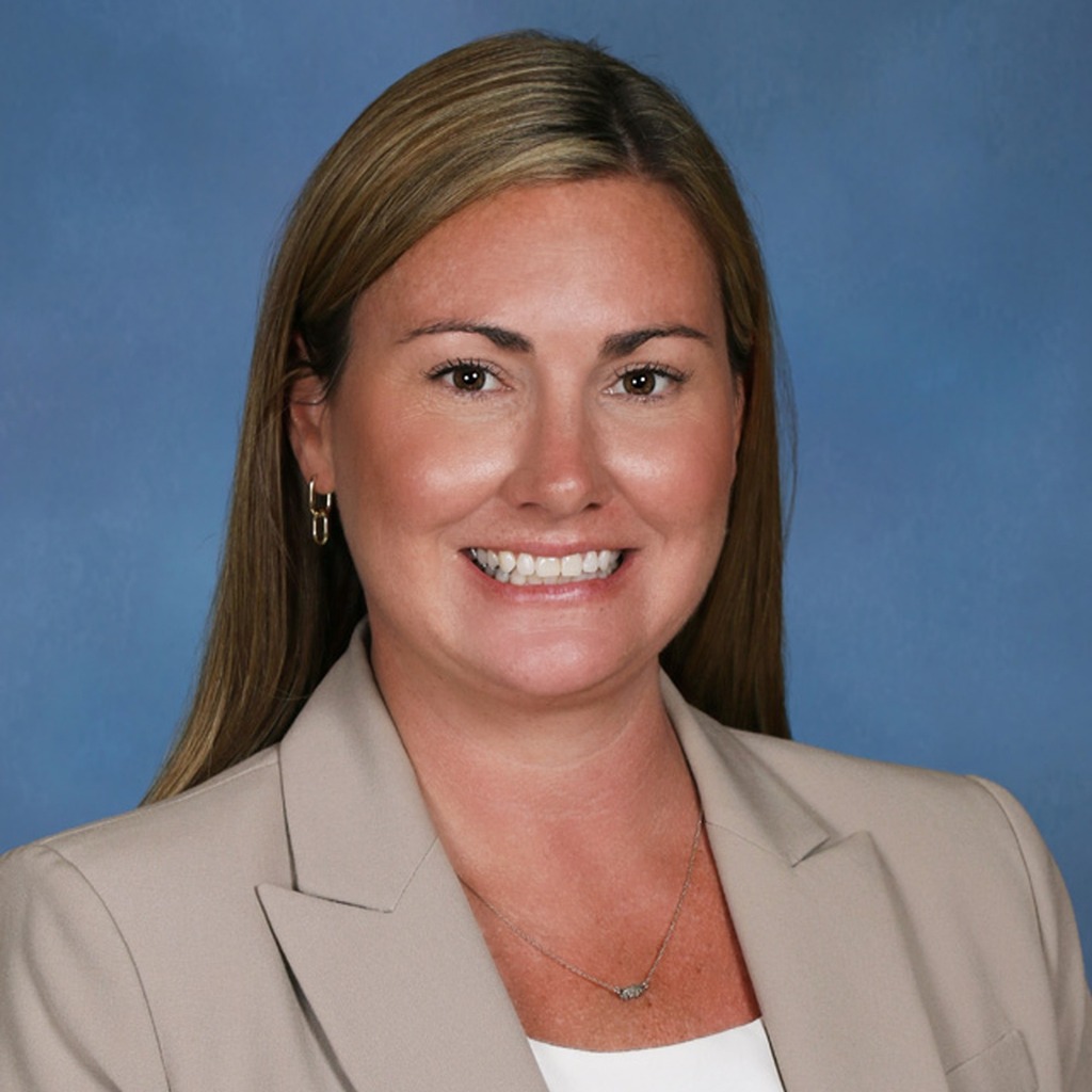 A woman in a light blazer, white shirt, and necklace smiles at the camera against a blue background.