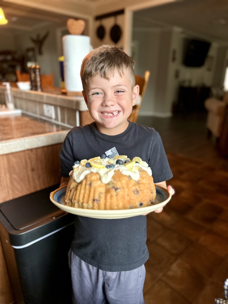 A boy holds a plate with a cake topped with blueberries. He is standing in a kitchen area.
