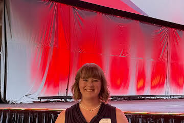 Person with short hair smiles in front of a stage with a red-lit backdrop and black curtains.