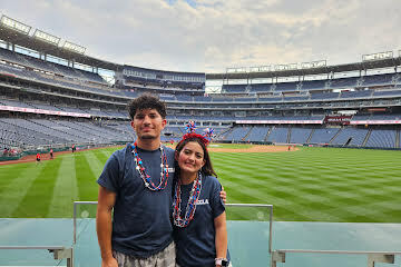 Two people stand in a stadium with empty seats, a wide baseball field, and a red, white, blue garland around their necks.