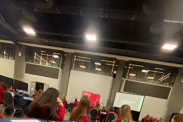 A room with many people seated at tables; a red banner on a stage, projector screen.