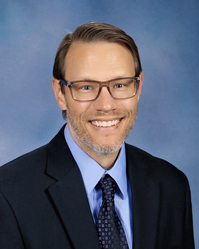 A man in a blue shirt and black suit with a tie and glasses. He is smiling.