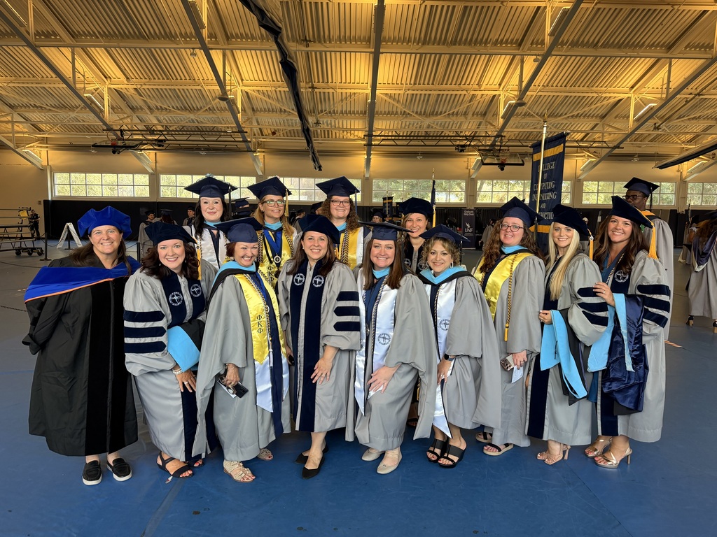 A group of people in academic regalia, including caps and gowns, stand in a large indoor space.