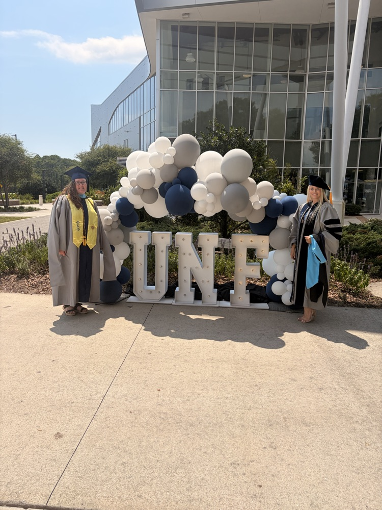 Mrs. Thomas and Principal Metz standing in front of a sign that say UNF in their cap and gown