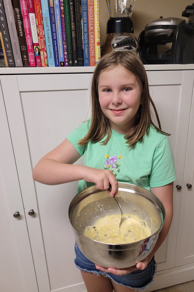 A young girl holding a mixing bowl filled with batter, standing in a room with white cabinets.