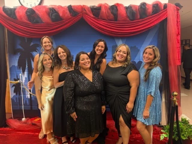 Eight women in evening wear, smiling, stand in front of a stage backdrop with a red curtain.