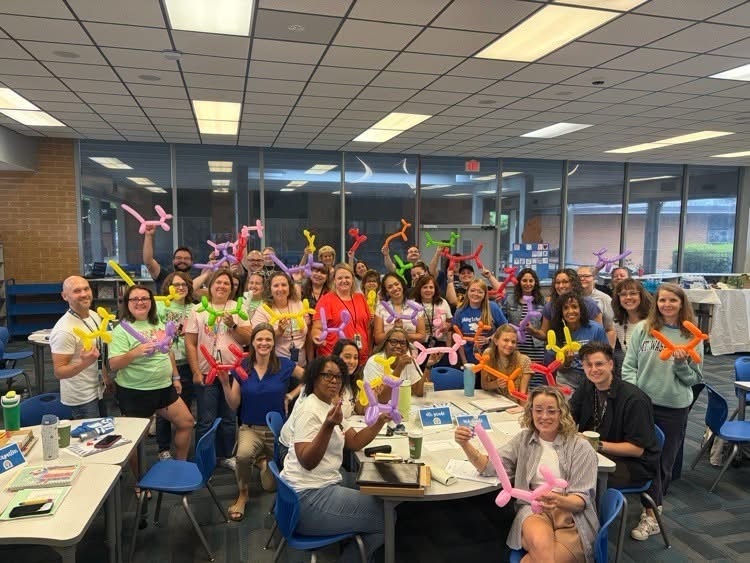 Group photo of people with balloon animals in a room with tables, chairs, and large windows.