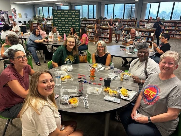 A group of adults in a library setting sitting around tables with food, drinks, and various items.
