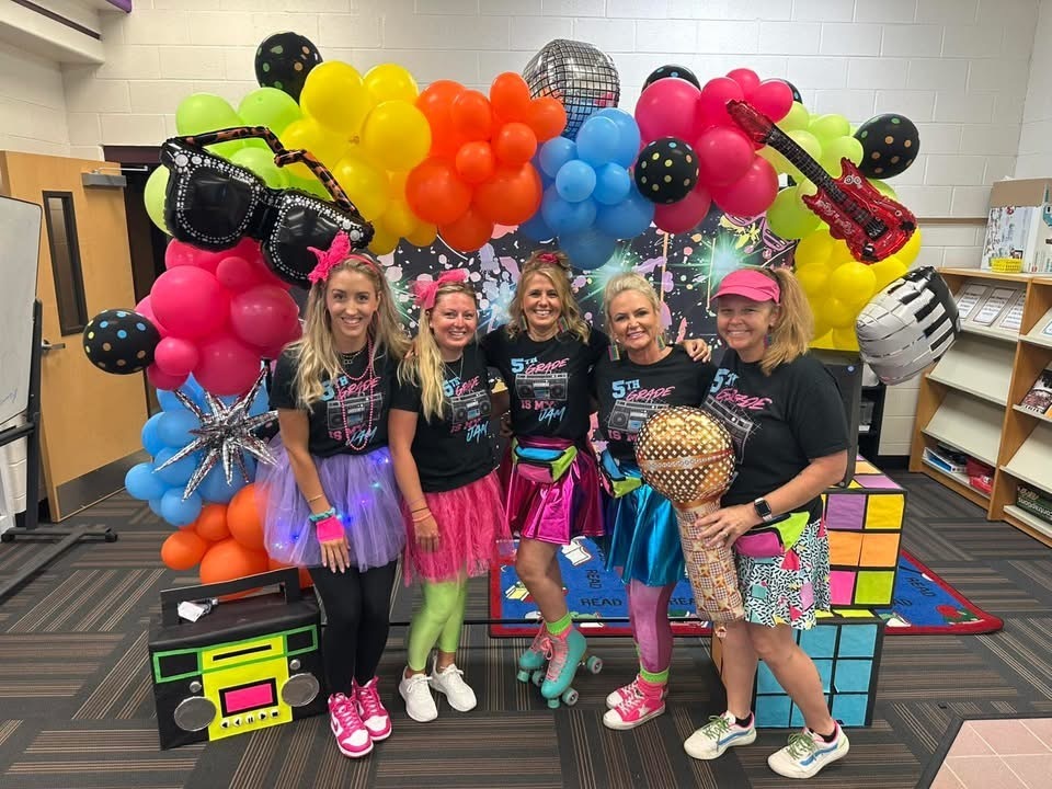 Five women in 80s-themed outfits pose before a colorful balloon arch, Rubik's Cube, and a guitar.