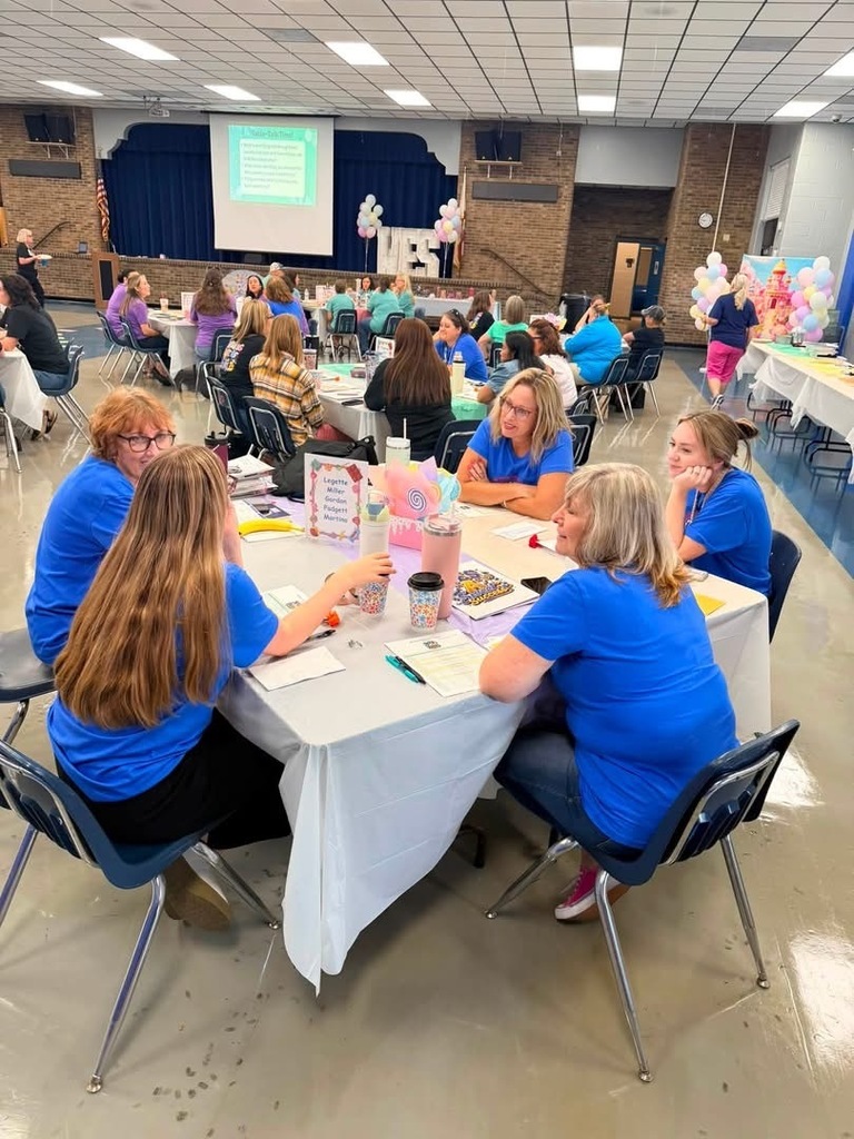 Several women in blue shirts sit at tables in a room with balloons. A projector screen is in the background.