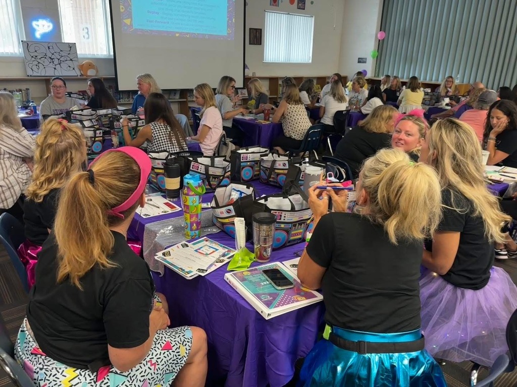 Group of women sitting at tables with purple tablecloths, engaged in an activity. A projector screen and whiteboard visible in background.