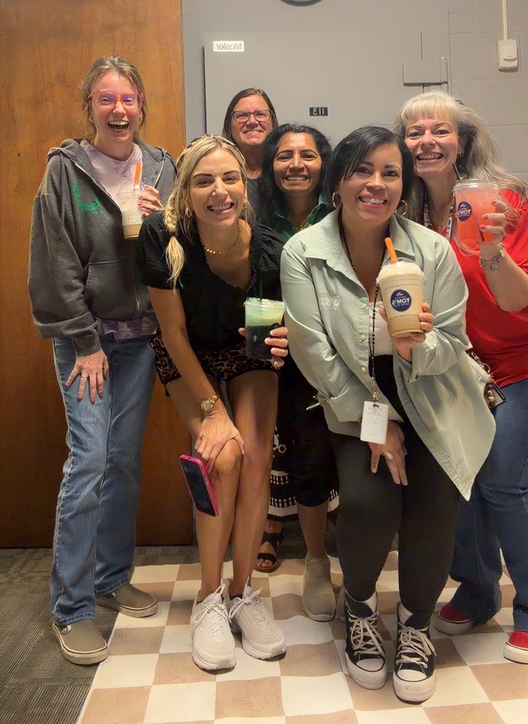 A group of smiling women poses for a photo in a room with checkered floor. They hold cups and one has a phone.